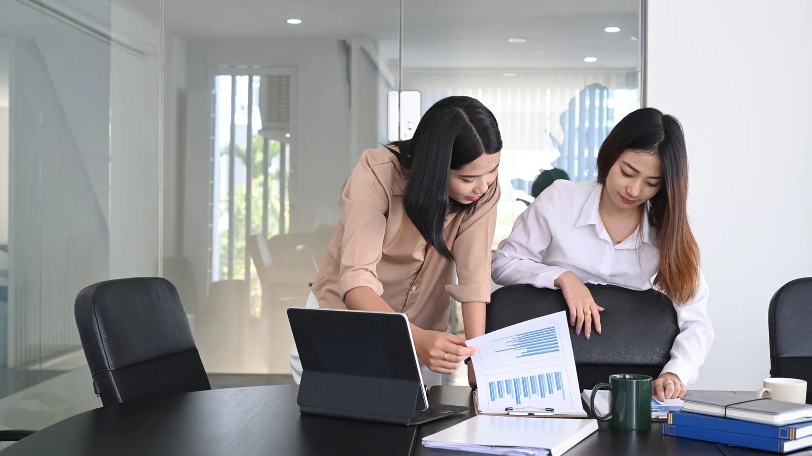 Two colleagues reviewing a printed chart beside a laptop in a glass-walled modern conference room