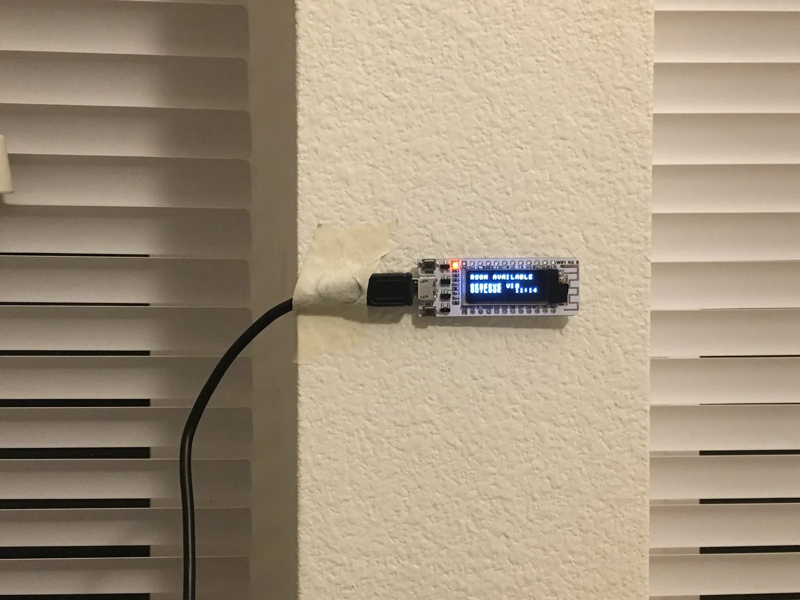 ESP8266 board with a small OLED screen taped beside a meeting room door showing Room Available
