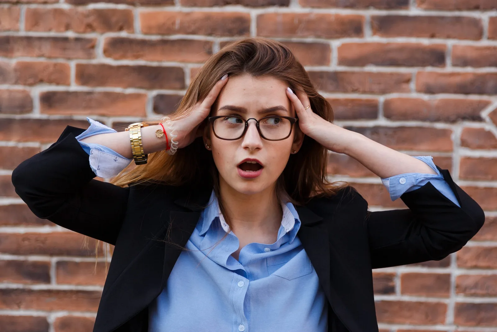 Person in a blazer looking shocked with hands to their head against a brick wall