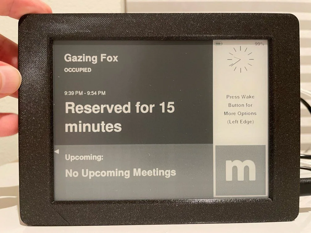 Hand holding an E-ink meeting room display showing the room reserved for 15 minutes