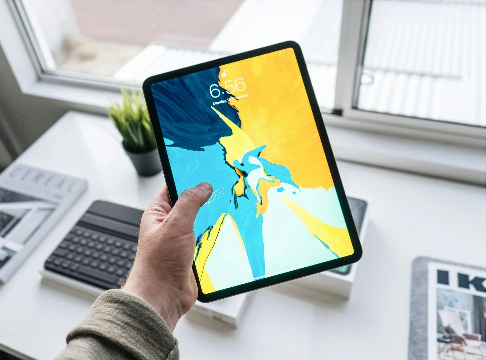 Hand holding an iPad Pro above a desk with a keyboard case and magazines