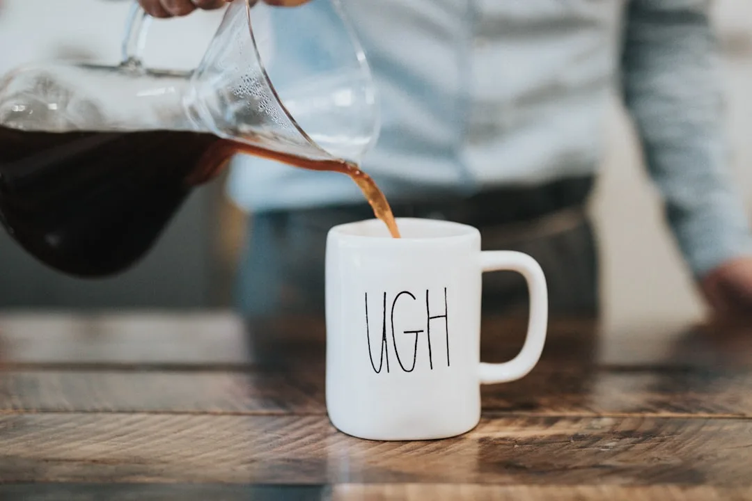Coffee being poured from a carafe into a mug printed with the word UGH