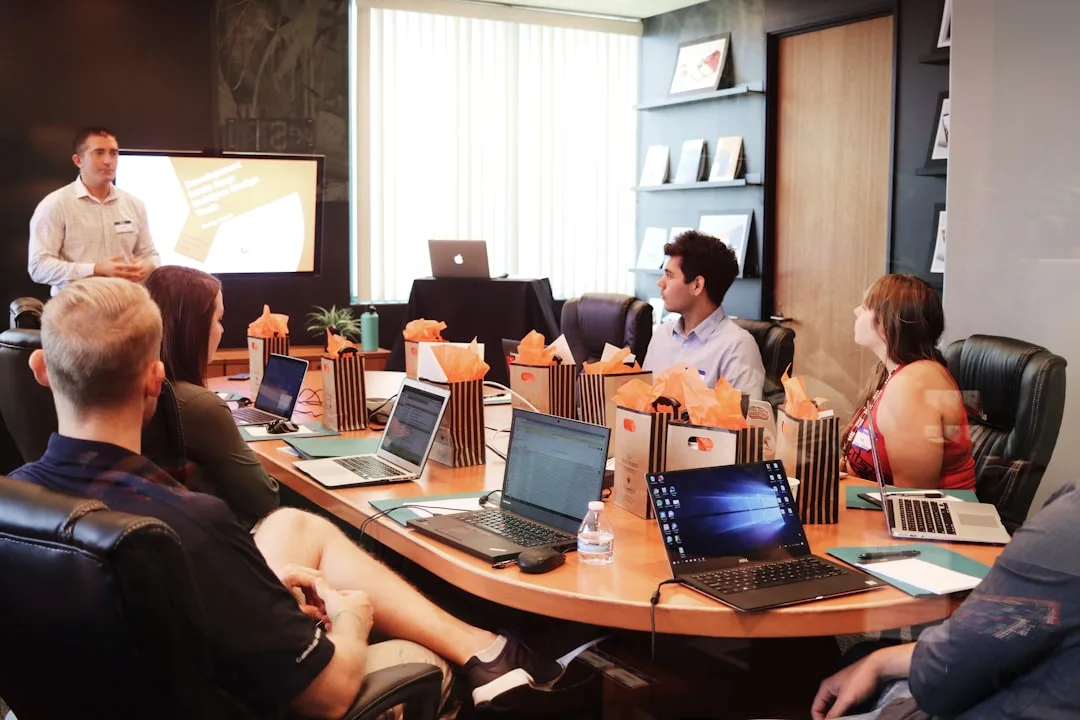 Presenter standing beside a screen addressing colleagues seated with laptops around a boardroom table